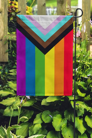 Progress Pride flag hanging from a garden pole placed in a garden. The Progress Pride Flag includes a black, brown, blue, pink and white stripe layered on top of the rainbow colors.