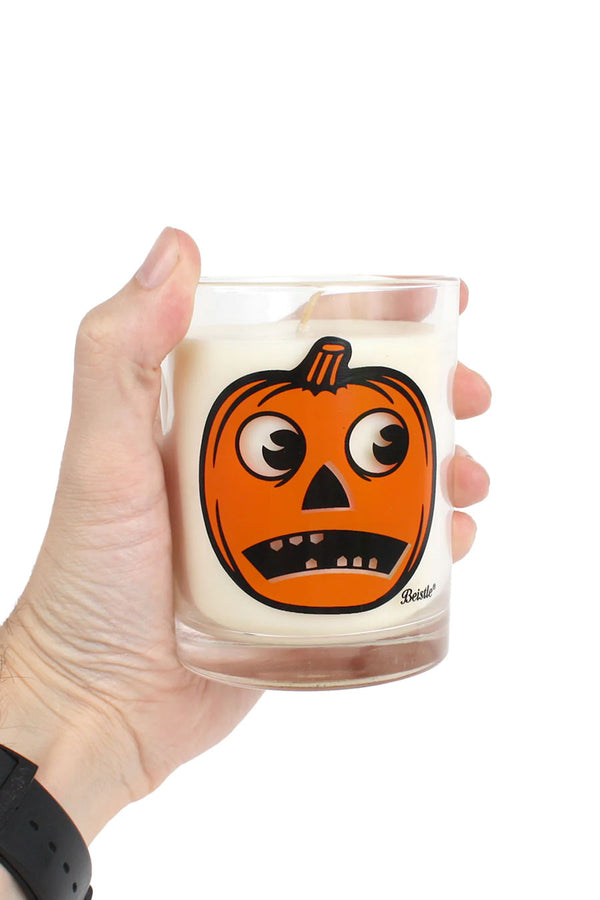 Person holding a scented candle in a clear jar. The jar features a Beistle jackolantern design. White background.