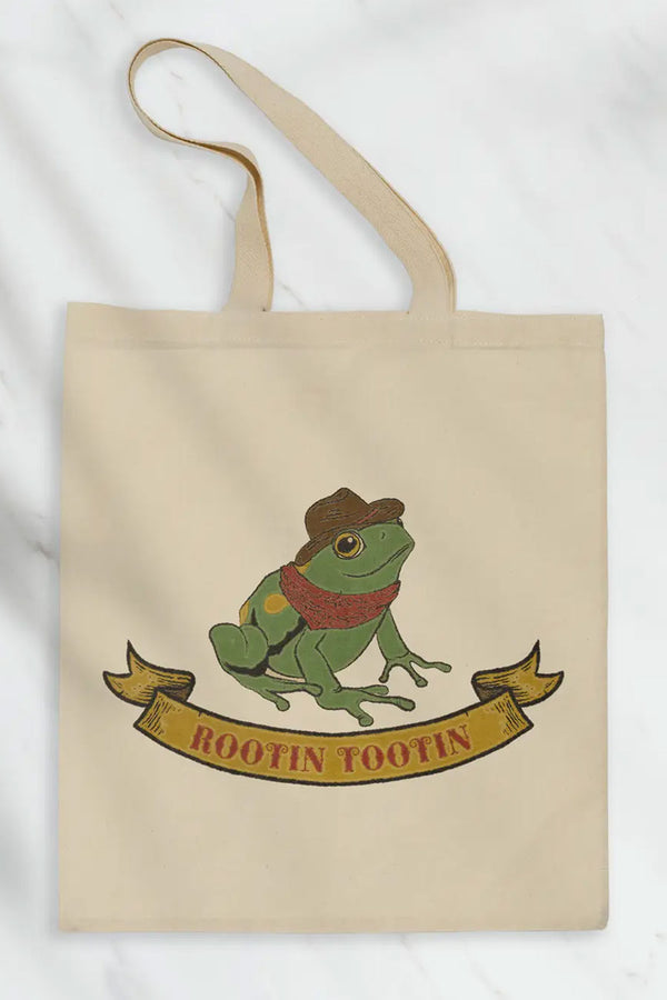 Natural cotton tote featuring illustration of a green frog wearing a red bandana and a cowboy hat. Under the frog is a yellow banner with red western text that says Rootin Tootin. White background.