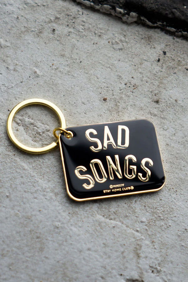 Keychain in hard black enamel and metal with epoxy coating and gold hardware. The keychain says Sad Songs in gold. Concrete background.