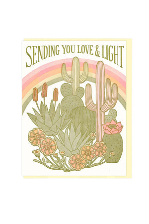 Greeting card with a rainbow across and cacti and desert flowers in front. Above the rainbow, the card says Sending you Love and Light. Yellow envelope. White background.