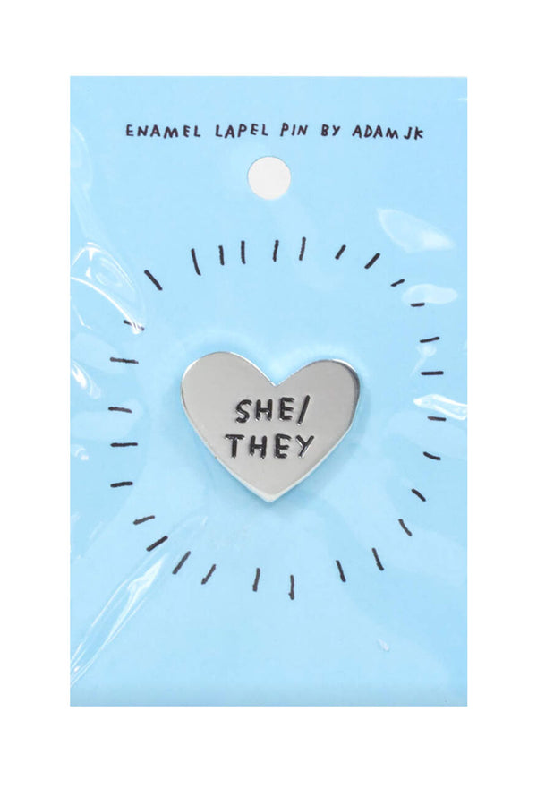 Silver heart shape enamel pin. The pin says She/They in black text. Pin is on a blue backing board. White background.