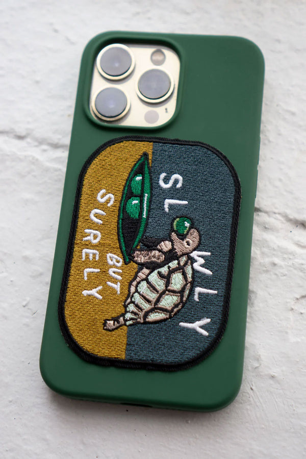 A cellphone with a green case featuring a A rounded rectangle patch that features a small turtle eating a snow pea with a yellow and blue background. The patch says Slowly But Surely in White text.