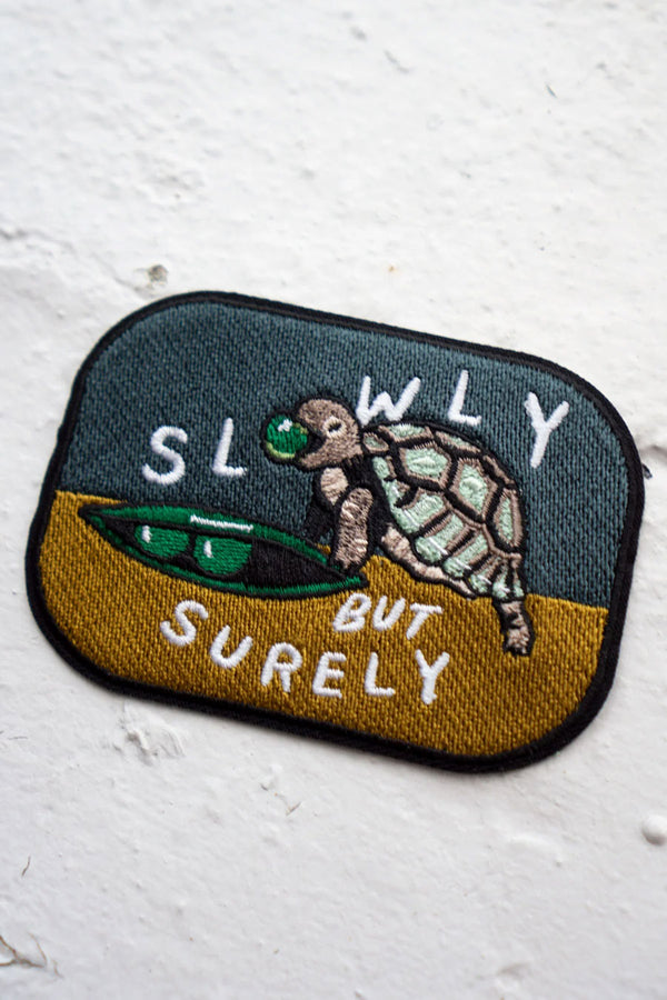 A rounded rectangle patch that features a small turtle eating a snow pea with a yellow and blue background. The patch says Slowly But Surely in White text. White background.