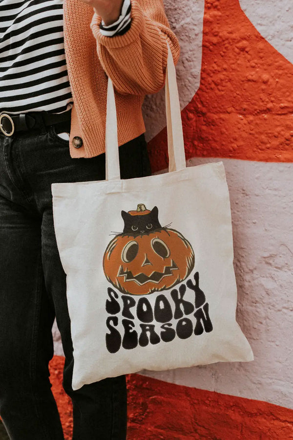 Person holding natural cotton tote bag featuring a jackolantern with a black kitten inside peeking its head out of the top of the pumpkin. Under the pumpkin the tote says Spooky season in large black bubbly letters.