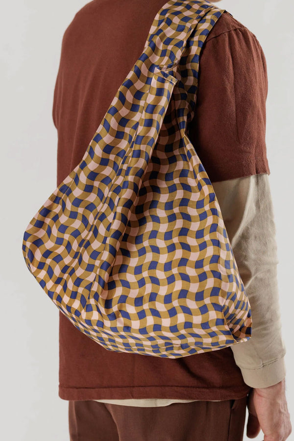 A person holding a Large reusable shopping back featuring a wavy gingham pattern in navy, peach, and gold. White background.