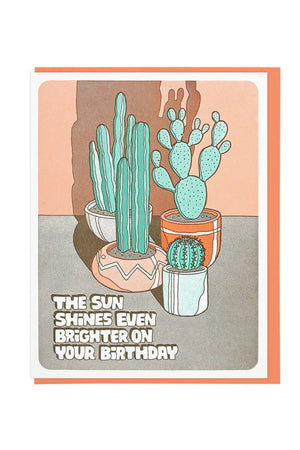 Greeting card featuring four different potted cacti in front of a peach wall. The card says The Sun shines even brighter on your birthday in block letters. Orange envelope. White background.