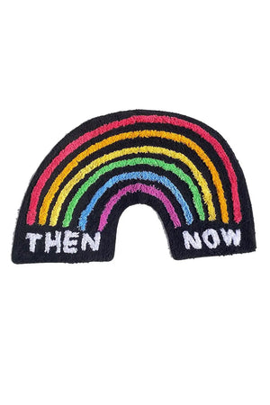 Black rainbow shaped rug featuring rainbow color striped arches. On the left side under the stripes the rug says Then and on the right at the end of the stripes the rug says Now. White background.