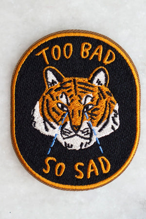 black oval patch with orange border. The patch features a crying orange tiger with the words "Too Bad So Sad" in orange text.