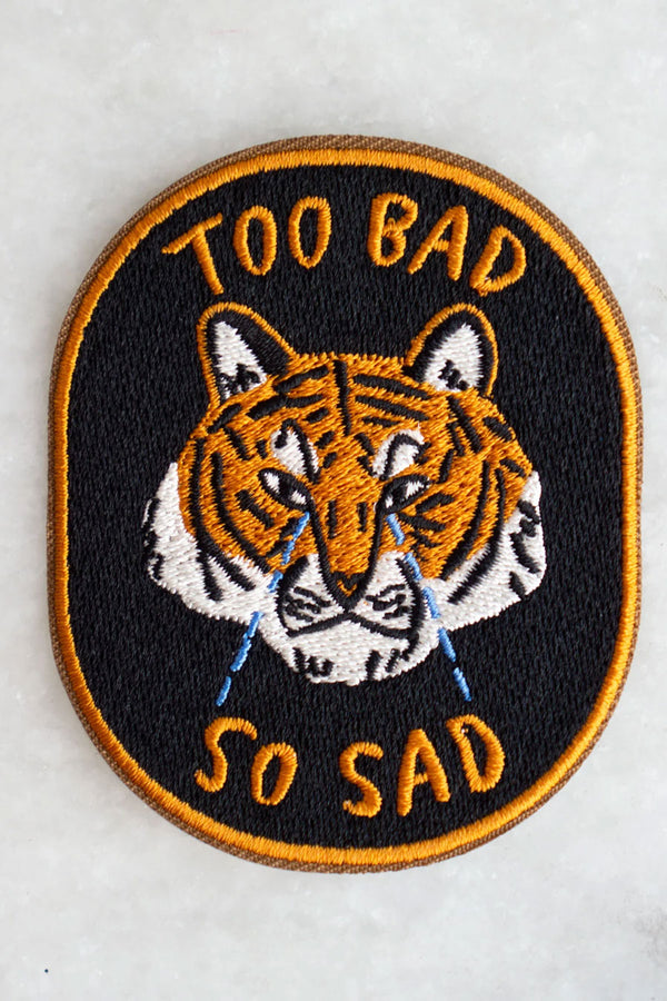 black oval patch with orange border. The patch features a crying orange tiger with the words "Too Bad So Sad" in orange text.