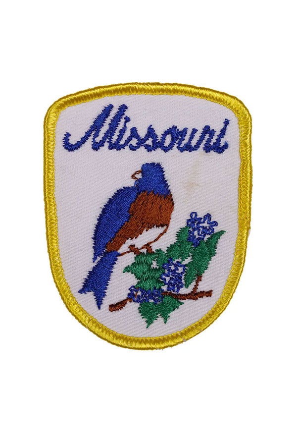 White patch with yellow border. The patch says Missouri in Blue cursive lettering and features a Bluebird sitting on a small branch with blue flowers. White background.