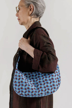 A person holding a Nylon crescent shaped bag featuring a wavy gingham pattern in blue, light blue, and red. Black straps. white background.