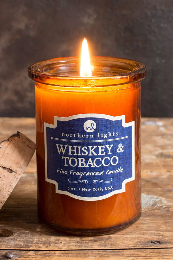 Amber glass candle with raw edge cork top. Blue label that says Whiskey and Tobacco. Natural wood backdrop.