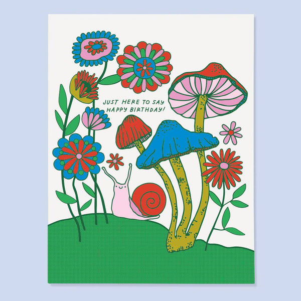 Birthday card featured illustrated mushrooms and flowers with a snail on green grass below them.  Limited color palette of pink, red, greens, and blue. The card says Just here to say Happy Birthday!