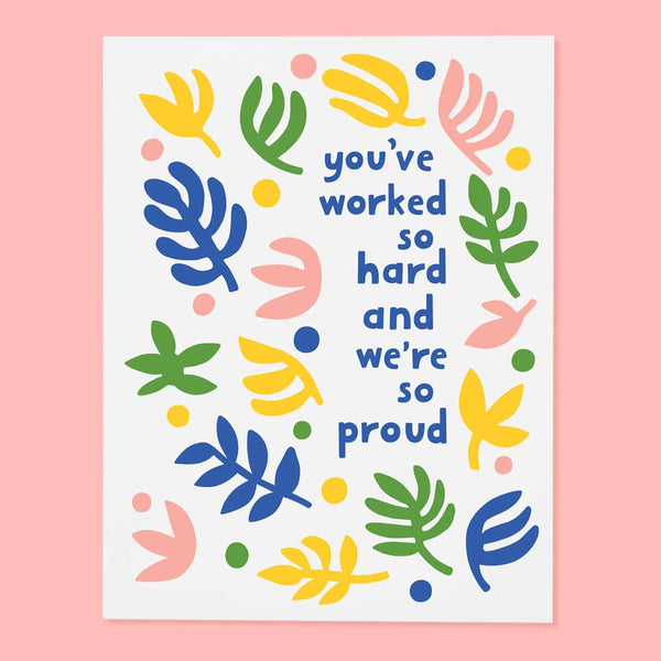 White greeting card  that says You've worked so hard and we're so proud. The text is surrounded by illustrations of plants and flowers in solid shapes in blue, green, pink, and yellow. Pink background.