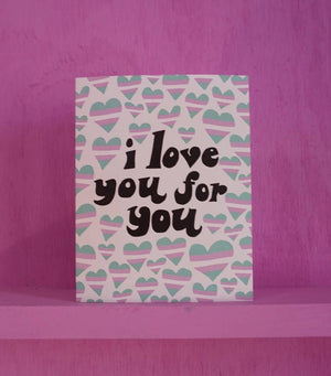 A pale pink greeting card with hearts in the color/pattern of the transgender flag. The card's text reads "I love you for you." Card is propped on a pink shelf with a pink wall in the background.