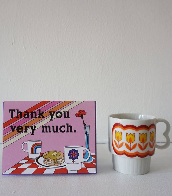 Pink Card with purple envelope on white backdrop.  The card says Thank you very much above a breakfast setting on a red and white checkered table cloth. On the table is a couple of coffee mugs, a stack of pancakes, and a carnation in a vase. The card is sitting on a white table in front a white wall next to a white coffee mug with yellow flowers surrounded by a red and orange loopy border.