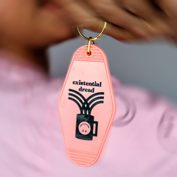 An image of a hand holding a pale pink keychain that says "Existential dread" in black lettering with an illustration of a coffee cup with an upside down smiley face.