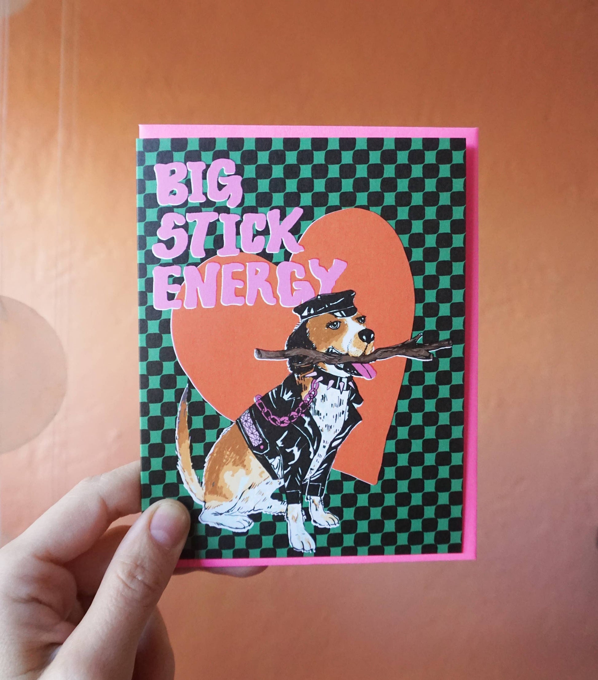 A green and black checkerboard patterned greeting card depicting a dog dressed in a leather outfit holding a stick. The text reads "Big stick energy" in pink letters.