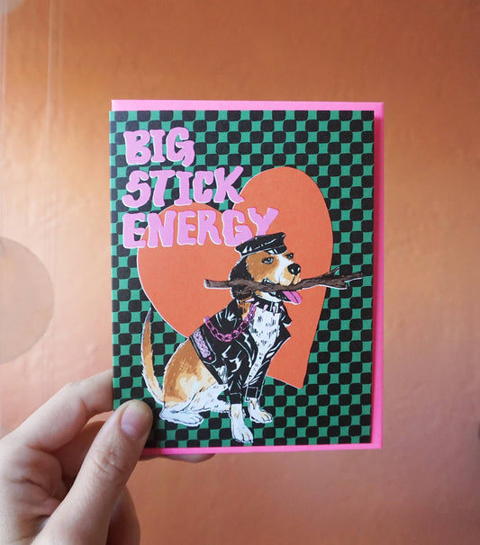 A green and black checkerboard patterned greeting card depicting a dog dressed in a leather outfit holding a stick. The text reads "Big stick energy" in pink letters.
