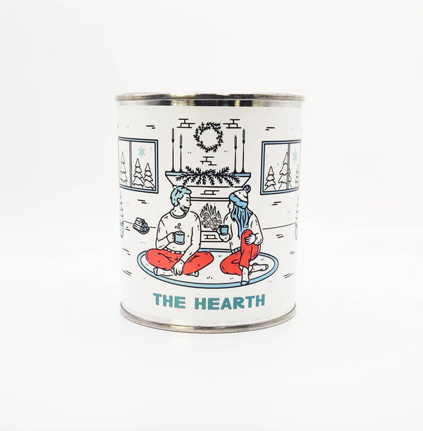 Candle in tin container. The label is white and features an illustration of two people drinking hot beverages in front of a fireplace. 