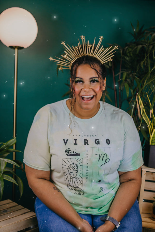 An olive-skinned woman is wearing a gold star crown and a lime gree, light blue, and yellow tie-dye Virgo t-shirt and jeans. The background has assorted plants, a dark green starry background and a gold floor lamp with a round globe light. 