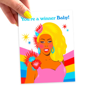 A hand is holding the corner of a greeting card with an illustration of Ru Paul in a blonde wig, a pink dress with red and blue flowers on the shoulder. The background has a pink, red, yellow, teal, and blue rainbow and the text reads "You're a winner Baby!" in blue.