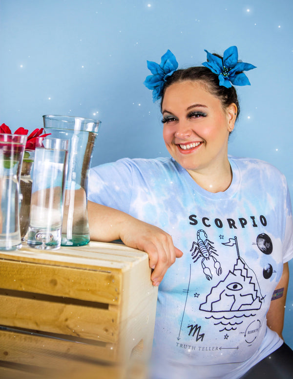 A dark-haired woman with 2 large blue flowers in her hair is wearing a blue, teal, and gray tie-dyed tshirt with the words "Scorpio." She is sitting next to a wooden crate with several glass vases full of water sitting on top. The shirt says "Scorpio" in black text along with the words "Truth teller" and "rebirth." It has illustrations of a scorpion, waves, planets, and a craggy mountaintop with an eyeball in the center and a small flag on top.