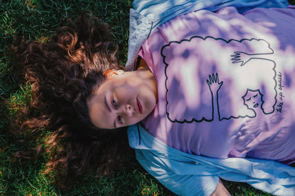 A Brunette woman laying on the grass wearing a blue button up shirt over a lilac tshirt. The shirt design is black line work of a person laying down with their hands above their head laying on their hair. The hair creates a rectangle border. Under the illustration the shirt says Take Up Space.