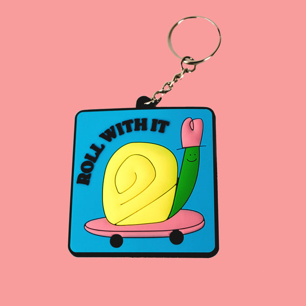 A square rubber keychain with a small silver chain and ring is sitting on a peach colored background. The keychain has a lightly textured image of a yellow and green snail riding a pink skateboard with black text that reads "roll with it."