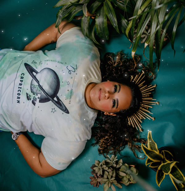 A photo of a olive-skinned woman with curly hair wearing a green and yellow tie-dyed shirt depicting symbols representing the astrological sign of "Capricorn."