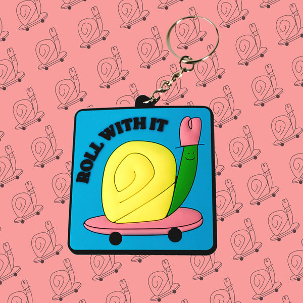 A square rubber keychain with a small silver chain and ring is sitting on a peach colored background with a pattern of black outlined snails on skateboards. The keychain has a lightly textured image of a yellow and green snail riding a pink skateboard with black text that reads "roll with it."