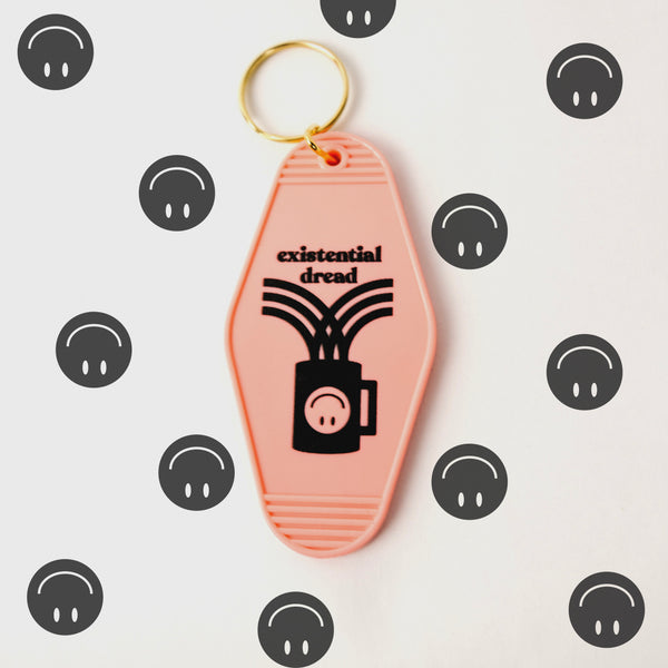 A photograph of a pale pink keychain with the words "existential dread" and a coffee cup with an upside down smiley face in black. The background is white with many upside down smiley faces.