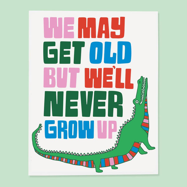 Greeting card that says We may get old but we'll never grow up. Below the text is a green alligator with a pink, red, and blue striped underbelly.