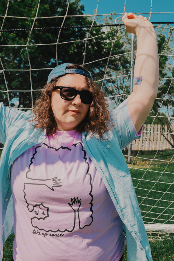 A Brunette woman standing in front of a soccer net wearing a blue baseball hat backwards, and button up shirt over a lilac tshirt. The shirt design is black line work of a person laying down with their hands above their head laying on their hair. The hair creates a rectangle border. Under the illustration the shirt says Take Up Space.