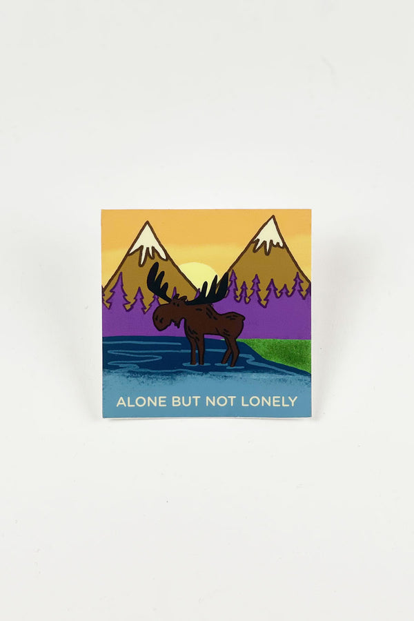 Sticker depicting a moose wading into water with. snow-covered peaks and a forest in the background. Sticker text says "Alone But Not Lonely."