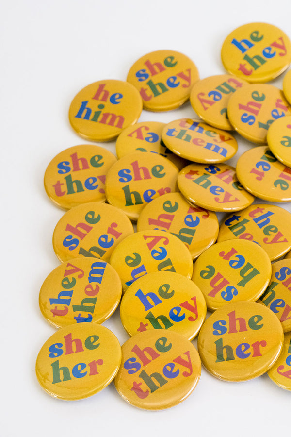 A pile of yellow pronoun buttons on a white background. The buttons say they them, she her, he him, she they, and he they. The colors of each letter alternates between blue, red, and green.