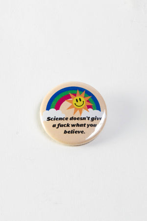 A round pinback button with a tan background, a rainbow stretched across two clouds, and a yellow and orange sun with a smiling face. The black text reads "Science doesn't give a fuck what you believe."