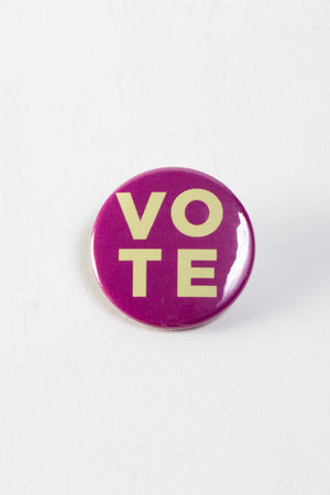 A magenta pinback button with cream colored text that says "VOTE"