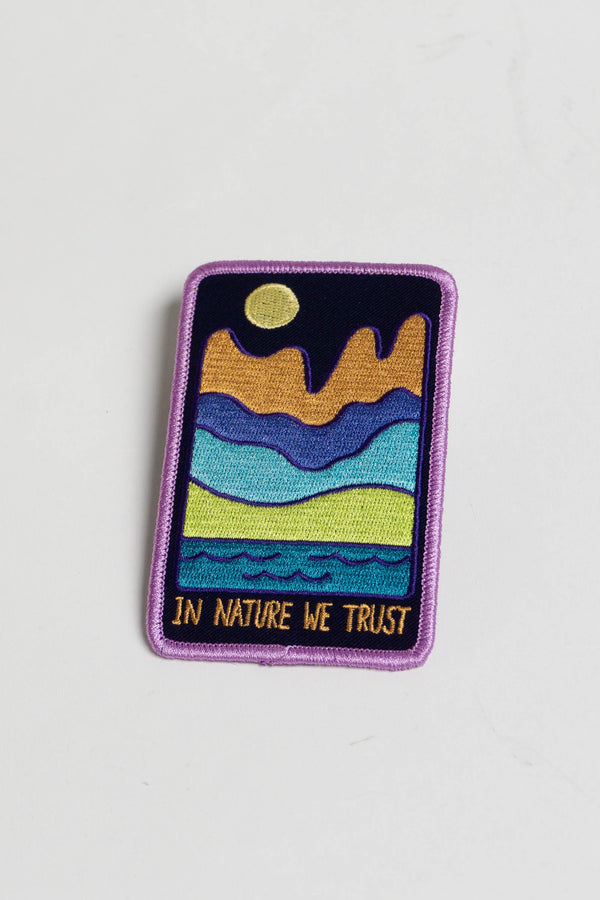 A dark blue rectangular patch depicting a mountainous landscape with a body of water in the foreground and a pale yellow moon in the sky. The text reads "In nature we trust" in orange. Patch has a lavender embroidered border.