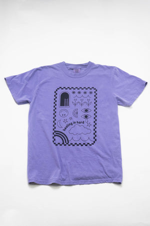 A purple t-shirt laying on a flat white background. The shirt's design is in purple ink with a checkerboard border and illustrations of clouds, smiley faces, tulips, stars, and rainbows. The text reads 