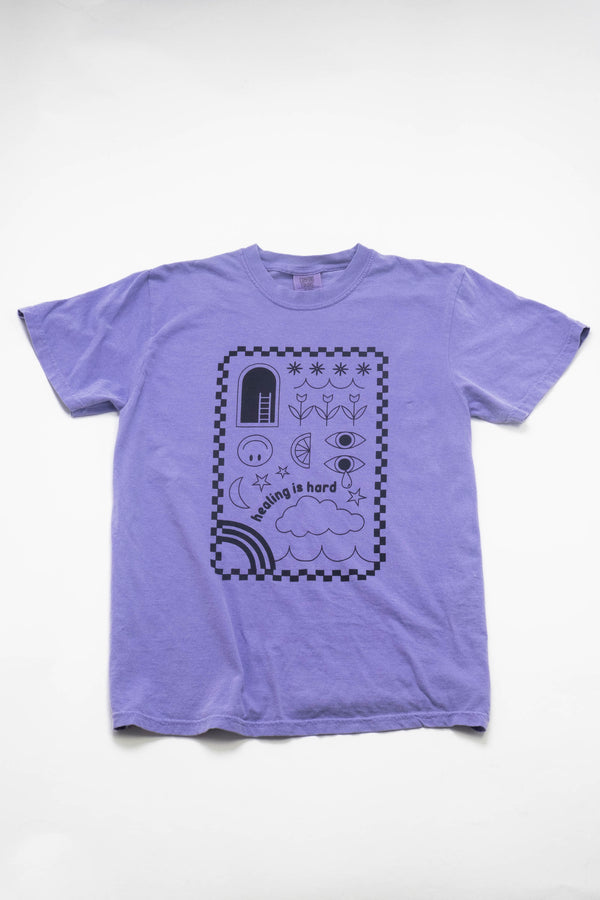 A purple t-shirt laying on a flat white background. The shirt's design is in purple ink with a checkerboard border and illustrations of clouds, smiley faces, tulips, stars, and rainbows. The text reads "healing is hard."