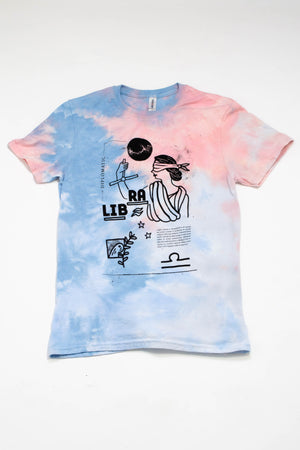 A light blue and pink tie-dyed tshirt on a white background. The shirt features black ink depictions of a blindfolded woman, a dove of peace, and small text describing the zodiac sign of Libra. The word "Libra" is prominently written in the center in block capital letters.