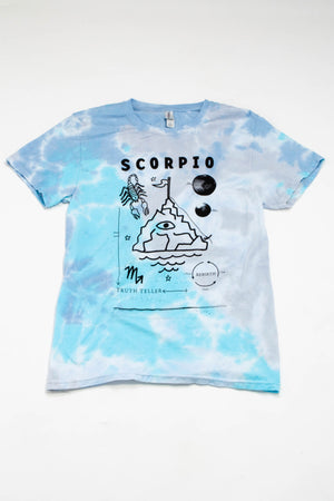 A blue, teal, and gray tie-dyed tee laying on a flat white background. The shirt says "Scorpio" in black text along with the words "Truth teller" and "rebirth." It has illustrations of a scorpion, waves, planets, and a craggy mountaintop with an eyeball in the center and a small flag on top.