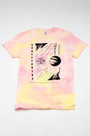 A yellow and pink tie-dye t-shirt lies on a flat white surface. The shirt is illustrated with symbols attributed to the zodiac sign Sagittarius and features text that reads "Sagittarius," "Generous," and "Adventurous."