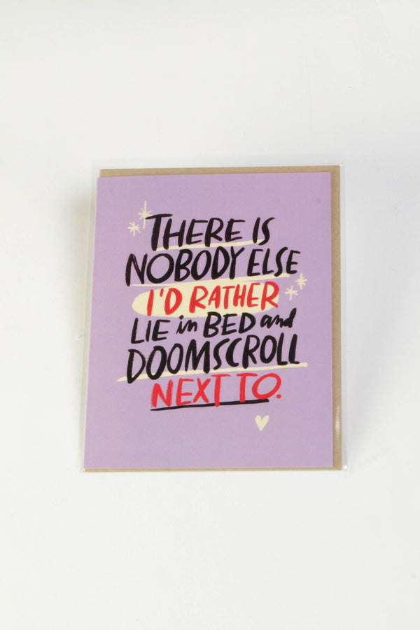 A lavender greeting card with black and red text that reads "There is nobody else I'd rather lie in bed and doomscroll next to."