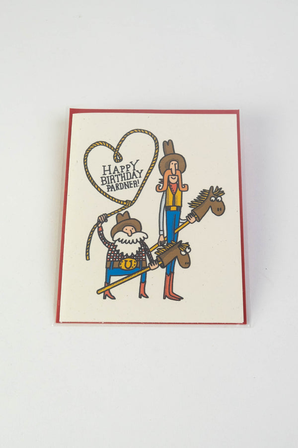 A white greeting card with a whimsical illustration of 2 cowboys riding stick horses. The short cowboy is swinging a lasso in the shape of a heart. The text reads "Happy Birthday Pardner!"