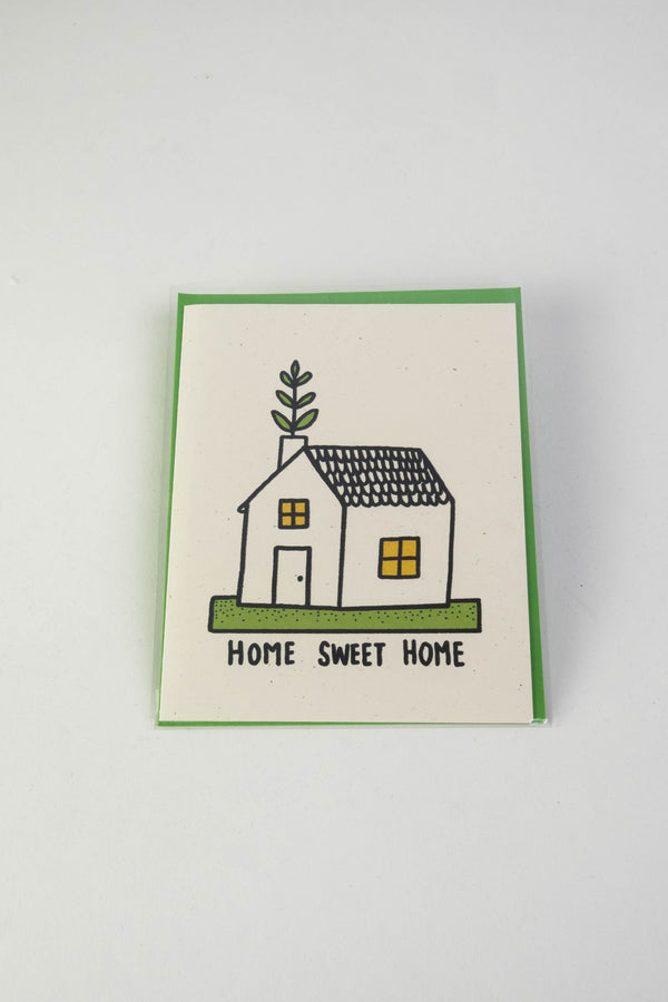 A white greeting card with the illustration of a small house with green foliage coming out of the chimney. The text along the bottom reads "Home sweet home."