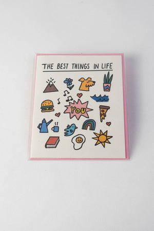 White card with pink envelope on a white backdrop. The card says The Best Things In Life with a line under neath. Below the line are small illustrations of a mountain, flower, dog, house plant, hamburger, waves, pizza, rainbow, sun, egg, blue bird, book, tea kettle and mug. In the center of the card is a pink starburst with the word YOU in bold yellow letters.