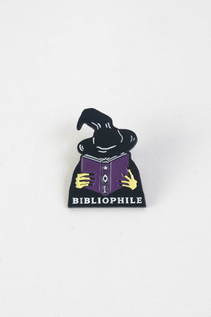 A die-cute enamel pin of a witch reading a book of spells. Text on the bottom of the pin reads "Bibliophile."